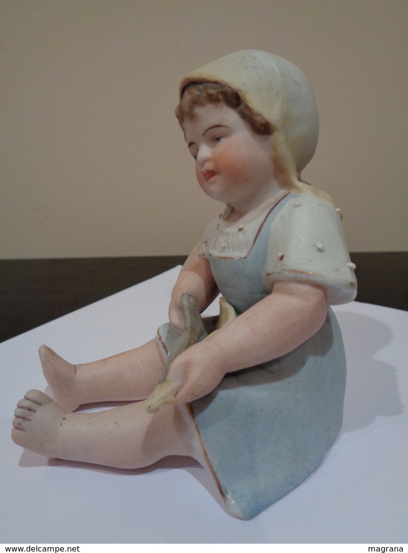 Muñeca de porcelana biscuit. Niña con gorro, sentada con unas tijeras.