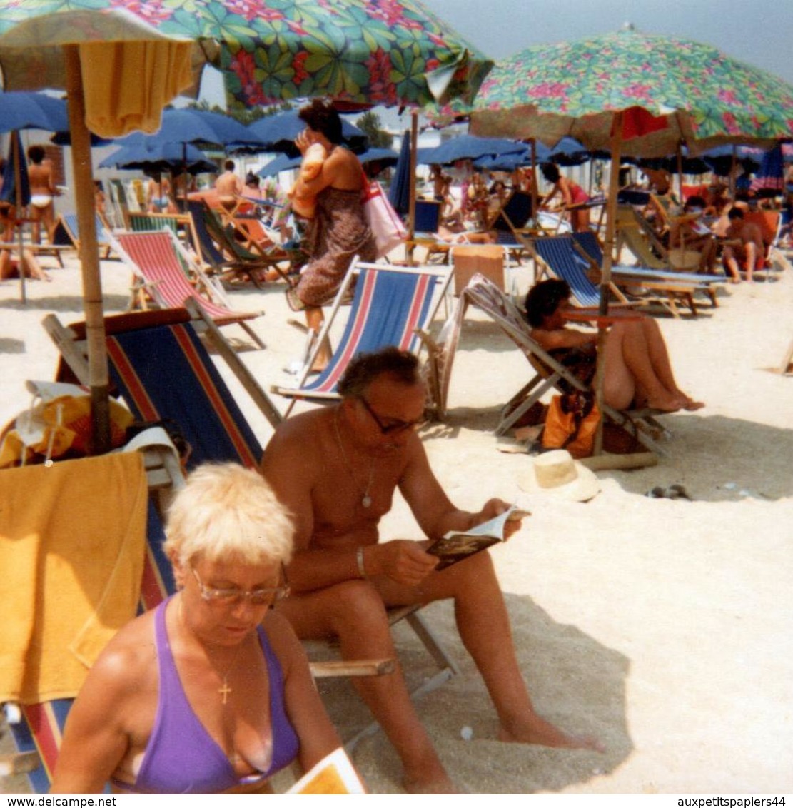 Photo Carrée Couleur Vintage 1980's - Couple De Retraités Pour Lecture Et Farniente En Maillots De Bains à La Plage - Personnes Anonymes