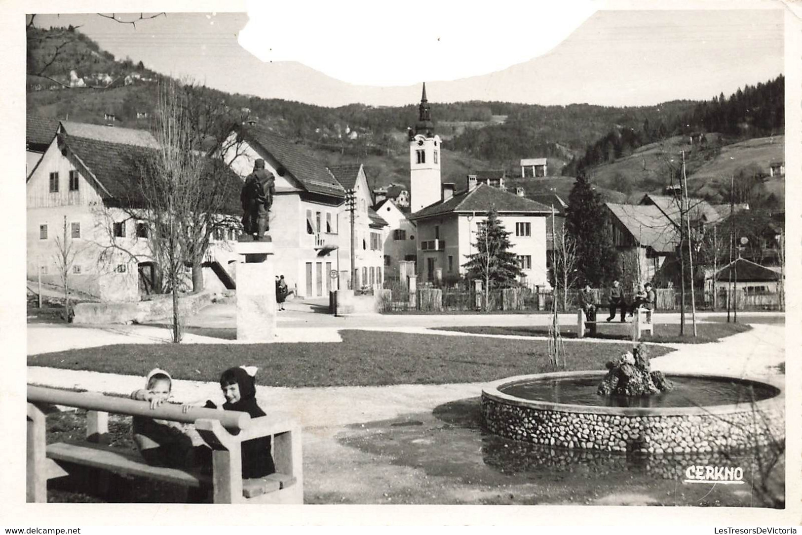 CP De Slovénie - Cerkno - Deux Enfants Sur Un Banc Devant Une Fontaine Et Un Village - Abimée Sur Le Haut Voir Scann - Slovenia