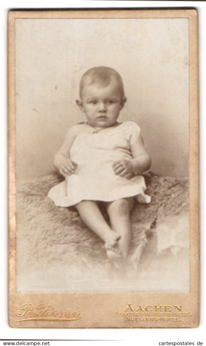 Fotografie R. Jarmer, Aachen, Friedrich-Wilhelm-Platz 5, Portrait Süsses Baby Im Weissen Kleidchen Auf Fell Sitzend  - Anonyme Personen