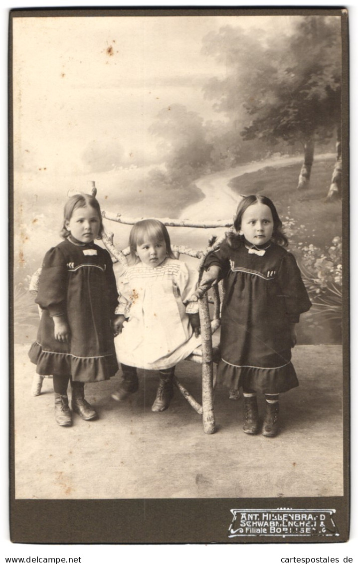 Fotografie Ant. Hillenbrand, Schwabmünchen, Drei Kleine Mädchen In Kleidern Auf Einer Holzbank Vor Einer Studiokulis  - Anonyme Personen
