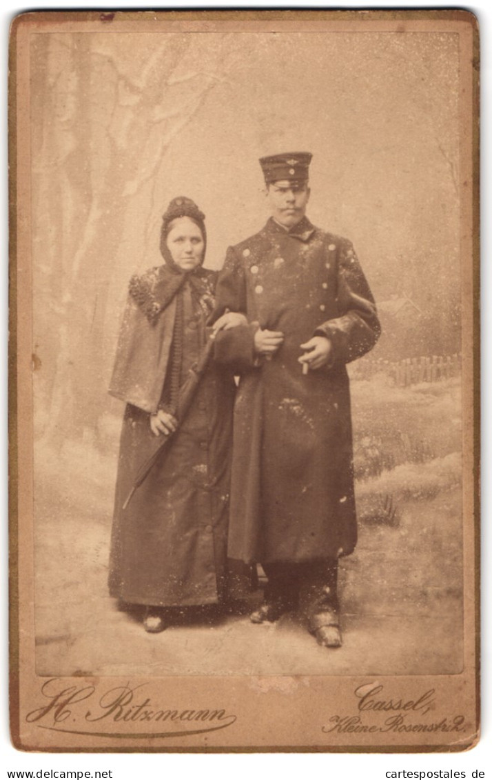 Fotografie H. Ritzmann, Cassel, Eisenbahner In Uniform Mit Mantel Und Mütze Nebst Frau In Einer Winter Studiokulisse  - Personnes Anonymes