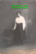 R407935 A Woman In A White Blouse And Black Skirts Is Standing By A Chair. Postc - Welt
