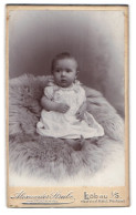 Fotografie Alexander Strube, Löbau I. S., Portrait Süsses Baby Im Weissen Kleidchen Auf Fell Sitzend  - Personnes Anonymes